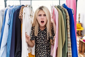 Naklejka na ściany i meble Young blonde woman searching clothes on clothing rack afraid and shocked with surprise and amazed expression, fear and excited face.