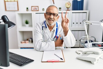 Mature doctor man at the clinic smiling with happy face winking at the camera doing victory sign....