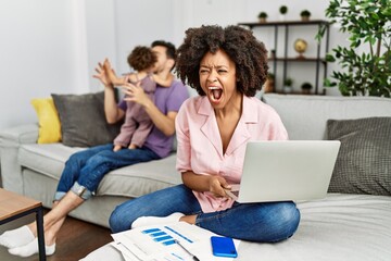 Mother of interracial family working using computer laptop at home angry and mad screaming frustrated and furious, shouting with anger. rage and aggressive concept.