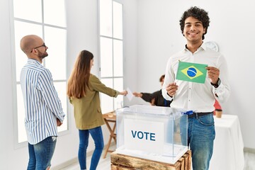 Young brazilian voter man smiling happy holding brazil flag standing by ballot at vote center.