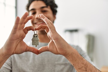 Young hispanic man doing heart symbol with hands holding key of new home.