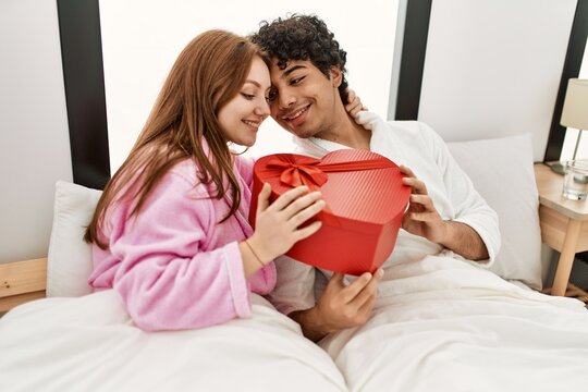 Young couple smiling happy holding valentine heart box gift at bedroom.