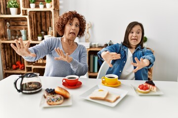 Family of mother and down syndrome daughter sitting at home eating breakfast afraid and terrified with fear expression stop gesture with hands, shouting in shock. panic concept.