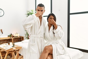 Young latin couple wearing towel standing at beauty center smelling something stinky and disgusting, intolerable smell, holding breath with fingers on nose. bad smell