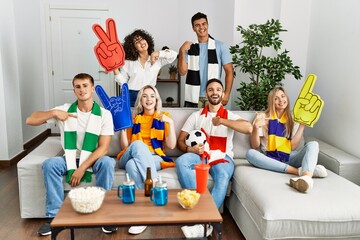 Group of friends supporting football team watching tv al home sitting on the sofa pointing finger...