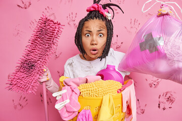 Domestic chores concept. Stunned young woman with braided hair holds dirty mop and bag of rubbish...