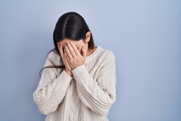 Young brunette woman standing over blue background with sad expression covering face with hands while crying. depression concept.