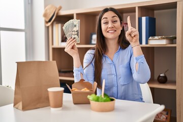 Young brunette woman eating take away food at home holding money smiling with an idea or question pointing finger with happy face, number one