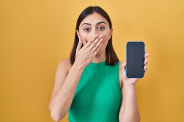 Hispanic girl holding smartphone showing screen covering mouth with hand, shocked and afraid for...