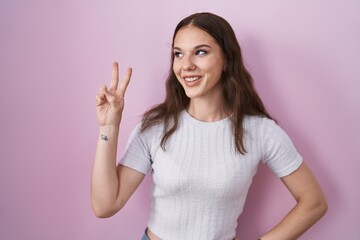 Young hispanic girl standing over pink background smiling looking to the camera showing fingers doing victory sign. number two.