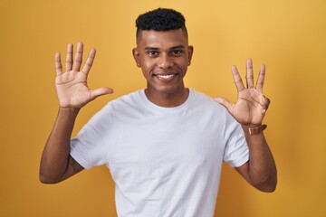 Young hispanic man standing over yellow background showing and pointing up with fingers number nine...