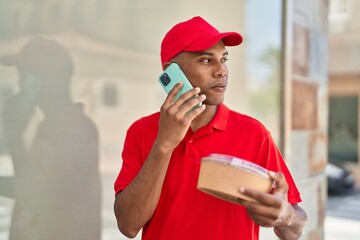 Young latin man delivery worker holding take away food talking on smartphone at street