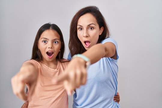 Young Mother And Daughter Standing Over White Background Pointing With Finger Surprised Ahead, Open Mouth Amazed Expression, Something On The Front