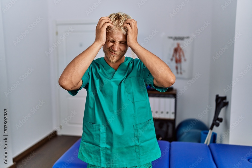 Sticker young blond man wearing physiotherapist uniform standing at clinic suffering from headache desperate