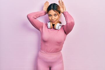 Beautiful hispanic woman wearing gym clothes and using headphones doing bunny ears gesture with...