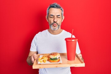 Handsome middle age man with grey hair eating a tasty classic burger with fries and soda looking at the camera blowing a kiss being lovely and sexy. love expression.