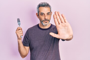 Handsome middle age man with grey hair holding one silver fork with open hand doing stop sign with...