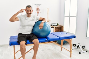 Middle age hispanic man at pain recovery clinic holding pilates ball smiling pointing to head with...