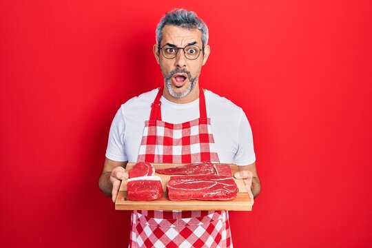 Handsome middle age man with grey hair wearing apron holding board with raw meat afraid and shocked with surprise and amazed expression, fear and excited face.