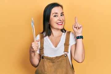 Middle age hispanic woman holding paintbrushes surprised with an idea or question pointing finger...