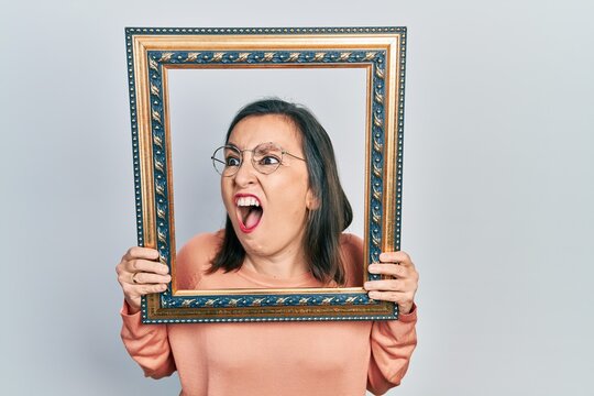 Middle age hispanic woman holding empty frame angry and mad screaming frustrated and furious, shouting with anger. rage and aggressive concept.