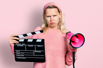 Young caucasian woman holding video film clapboard and megaphone skeptic and nervous, frowning...
