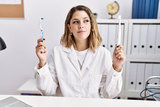 Young hispanic dentist woman holding electric toothbrush and teethbrush at clinic smiling looking to the side and staring away thinking.