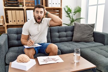 Handsome hispanic man working with rorschach test at psychology clinic crazy and scared with hands on head, afraid and surprised of shock with open mouth