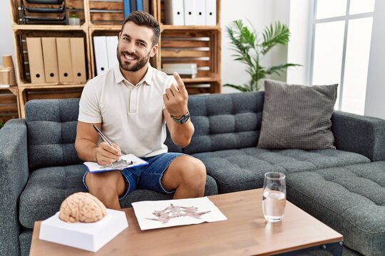 Handsome hispanic man working with rorschach test at psychology clinic beckoning come here gesture with hand inviting welcoming happy and smiling