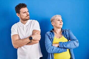 Young brazilian mother and son standing over blue background looking to the side with arms crossed convinced and confident