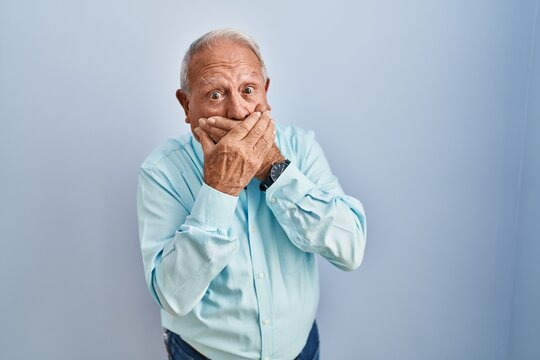Senior man with grey hair standing over blue background shocked covering mouth with hands for mistake. secret concept.