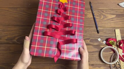 Women's hands demonstrate gift box with original design in Christmas style. Red ribbon is glued in form of fir tree and star made of orange peel on wrapper paper. Top view of Festive composition. - Powered by Adobe