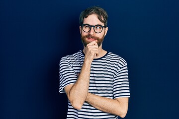 Caucasian man with beard wearing striped t shirt and glasses with hand on chin thinking about question, pensive expression. smiling with thoughtful face. doubt concept.