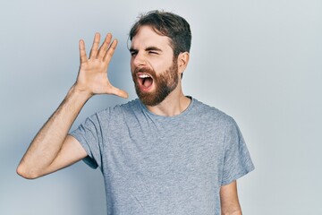 Caucasian man with beard wearing casual grey t shirt shouting and screaming loud to side with hand on mouth. communication concept.