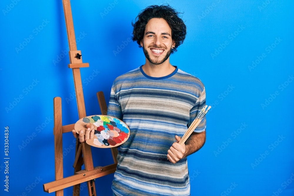 Poster handsome hispanic man standing drawing with palette by painter easel stand smiling with a happy and 