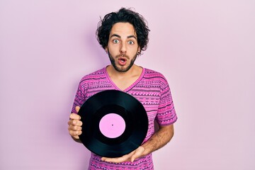 Handsome hispanic man holding vinyl disc afraid and shocked with surprise and amazed expression,...