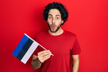 Handsome hispanic man holding estonia flag scared and amazed with open mouth for surprise,...