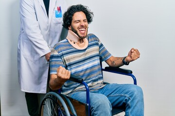 Handsome hispanic man sitting on wheelchair wearing neck collar very happy and excited doing winner gesture with arms raised, smiling and screaming for success. celebration concept.