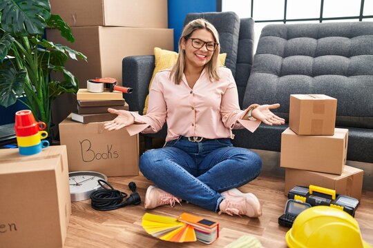 Young hispanic woman moving to a new home sitting on the floor smiling cheerful with open arms as friendly welcome, positive and confident greetings