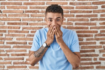 Brazilian young man standing over brick wall laughing and embarrassed giggle covering mouth with...