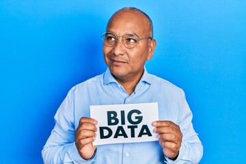 Middle age latin man holding paper with big data message smiling looking to the side and staring away thinking.