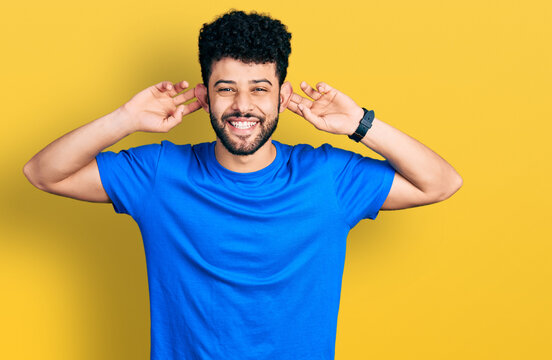 Young arab man with beard wearing casual blue t shirt smiling pulling ears with fingers, funny gesture. audition problem