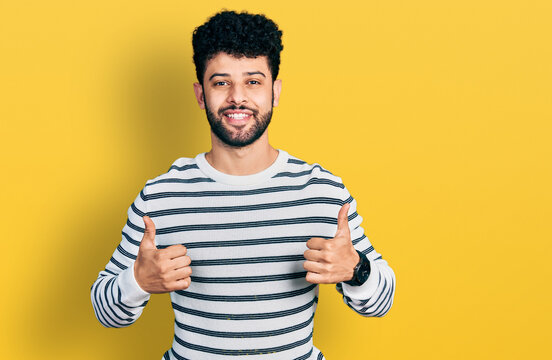 Young arab man with beard wearing casual striped sweater success sign doing positive gesture with hand, thumbs up smiling and happy. cheerful expression and winner gesture.