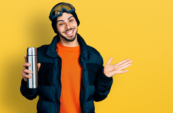 Young hispanic man with beard wearing snow wear and sky glasses holding thermo celebrating achievement with happy smile and winner expression with raised hand