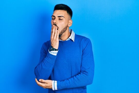 Young hispanic man with beard wearing casual blue sweater bored yawning tired covering mouth with hand. restless and sleepiness.
