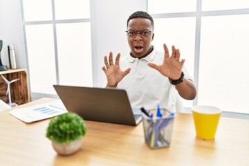 Young african man working at the office using computer laptop afraid and terrified with fear expression stop gesture with hands, shouting in shock. panic concept.