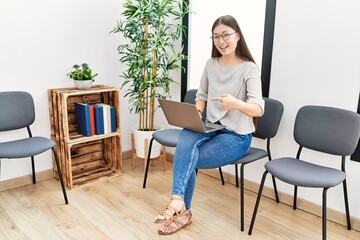 Young asian nurse woman sitting at waiting room using laptop smiling happy pointing with hand and finger