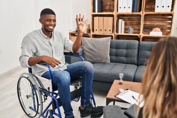 African american man doing therapy sitting on wheelchair showing and pointing up with fingers...