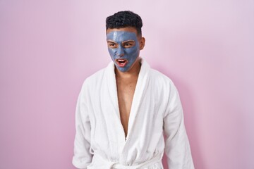 Young hispanic man wearing beauty face mask and bath robe angry and mad screaming frustrated and furious, shouting with anger. rage and aggressive concept.