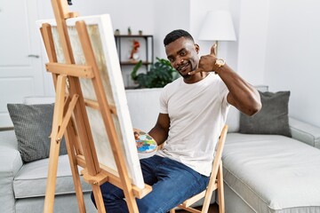 Young african man painting on canvas at home smiling doing phone gesture with hand and fingers like talking on the telephone. communicating concepts.